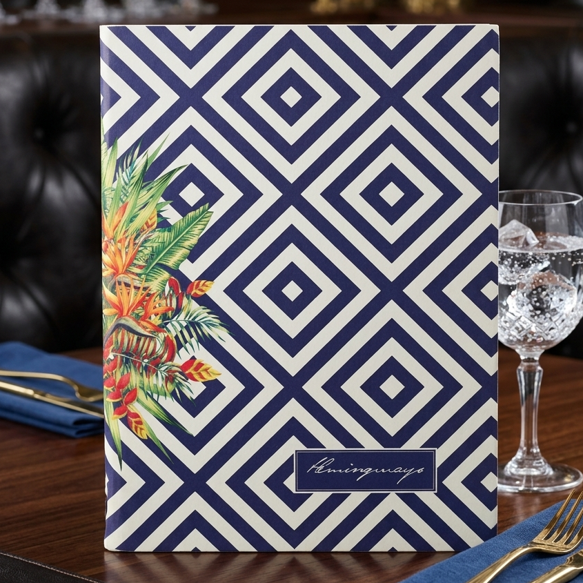 A rectangular menu or booklet standing on a wooden table, featuring a navy and cream geometric diamond pattern with a tropical floral illustration on the left and a small 'Huntingway' label near the bottom. A wine glass, blue napkins, and gold cutlery are arranged around it in a restaurant setting.