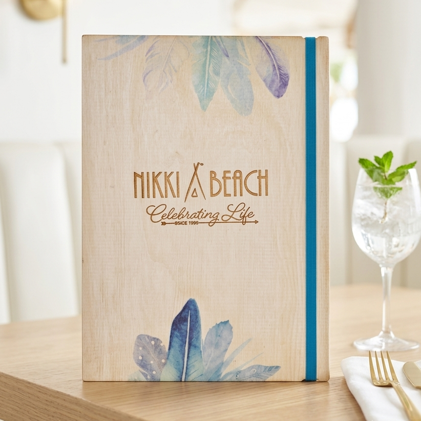 A personalized wooden menu or guest book standing on a light wood table in a bright dining setting. The cover is natural wood with blue and purple watercolor leaves at the top and bottom, and the text 'NIKKI & BEACH Celebrating Life' in brown.