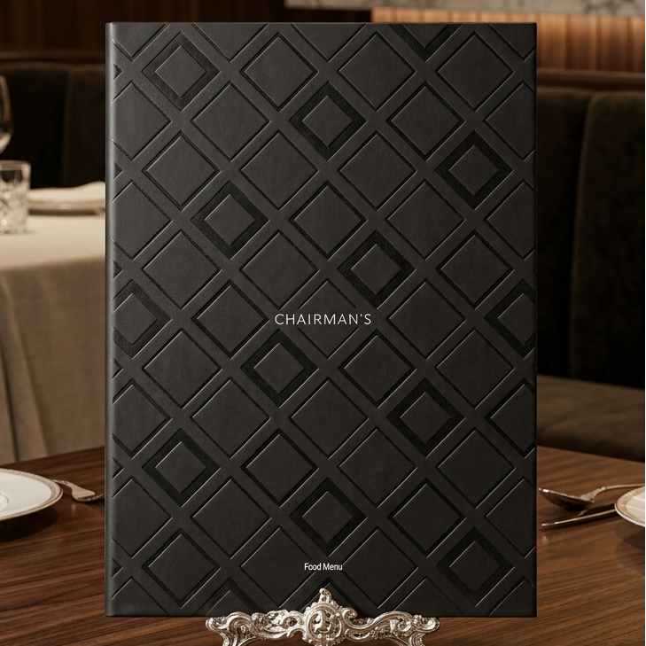 A black leather-bound menu or brochure with an embossed diamond pattern and the text 'CHAIRMAN'S' centered on the front, displayed on a small silver stand on a wooden dining table in a restaurant setting.
